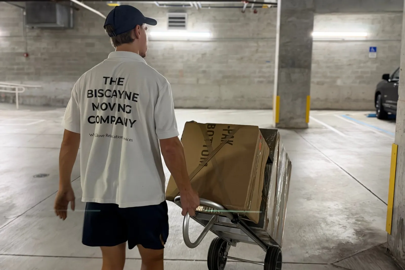 Biscayne Moving Co. mover wheeling a dolly of packed boxes through a Miami Beach condo parking garage